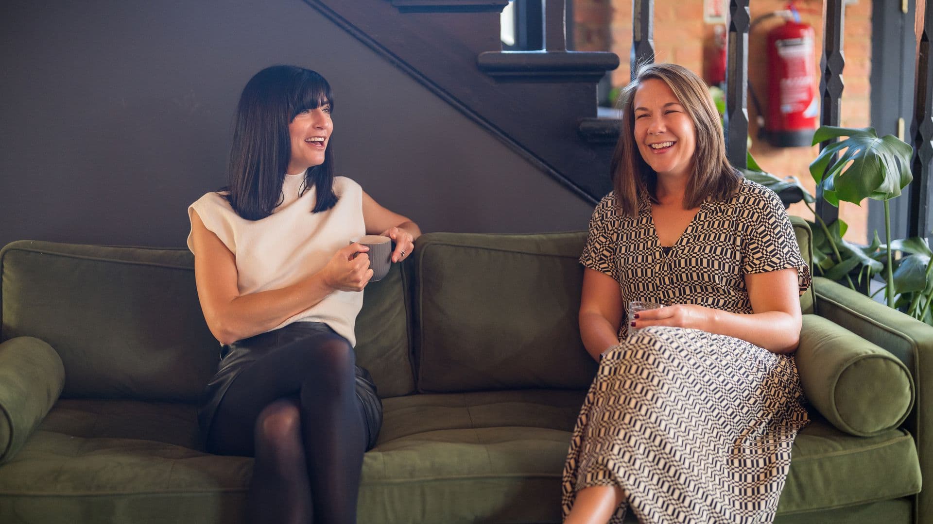 Katie Clarke and Leanne Terry laughing together on a green sofa, mugs in hand, on the filming day