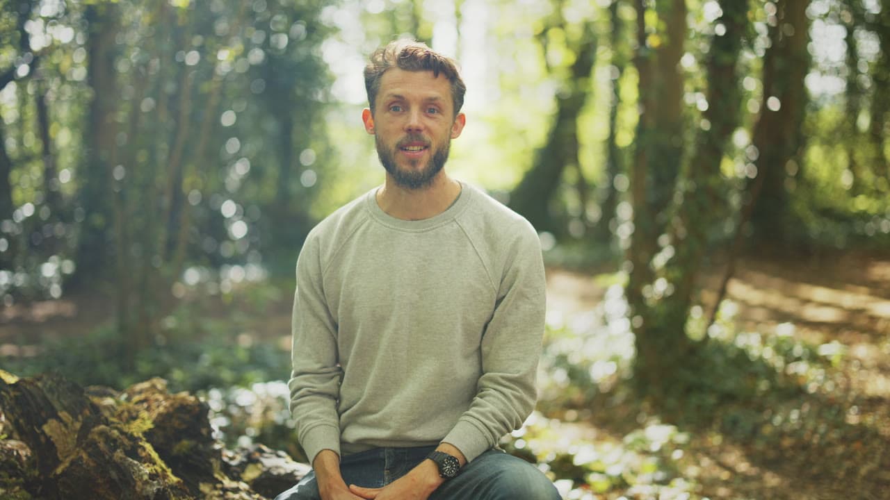 Jon sitting in a sunlit wood, smiling to the camera, in a short film
