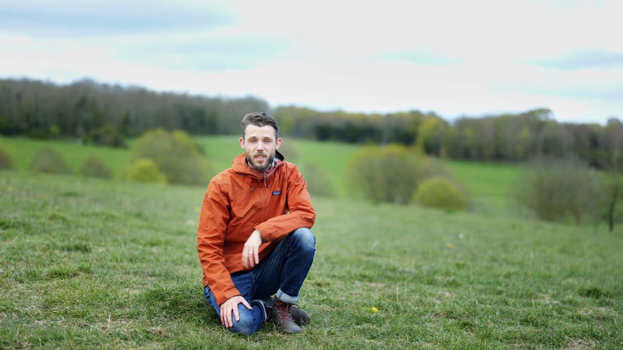 Jon in an orange jacket, crouched on the grass at Stanmer Park