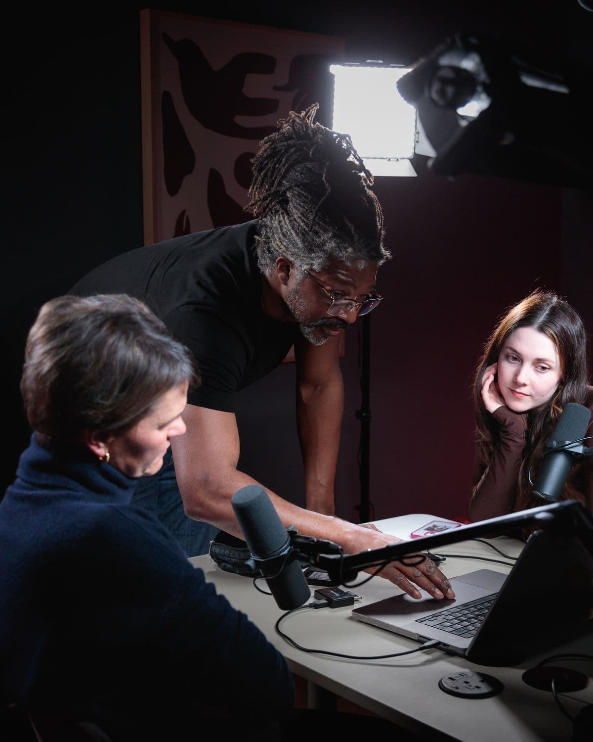 Kevan leaning over a laptop to play back the test recording to Mel Lawson and Liv Evans at the studio desk, under the set-up day's lights