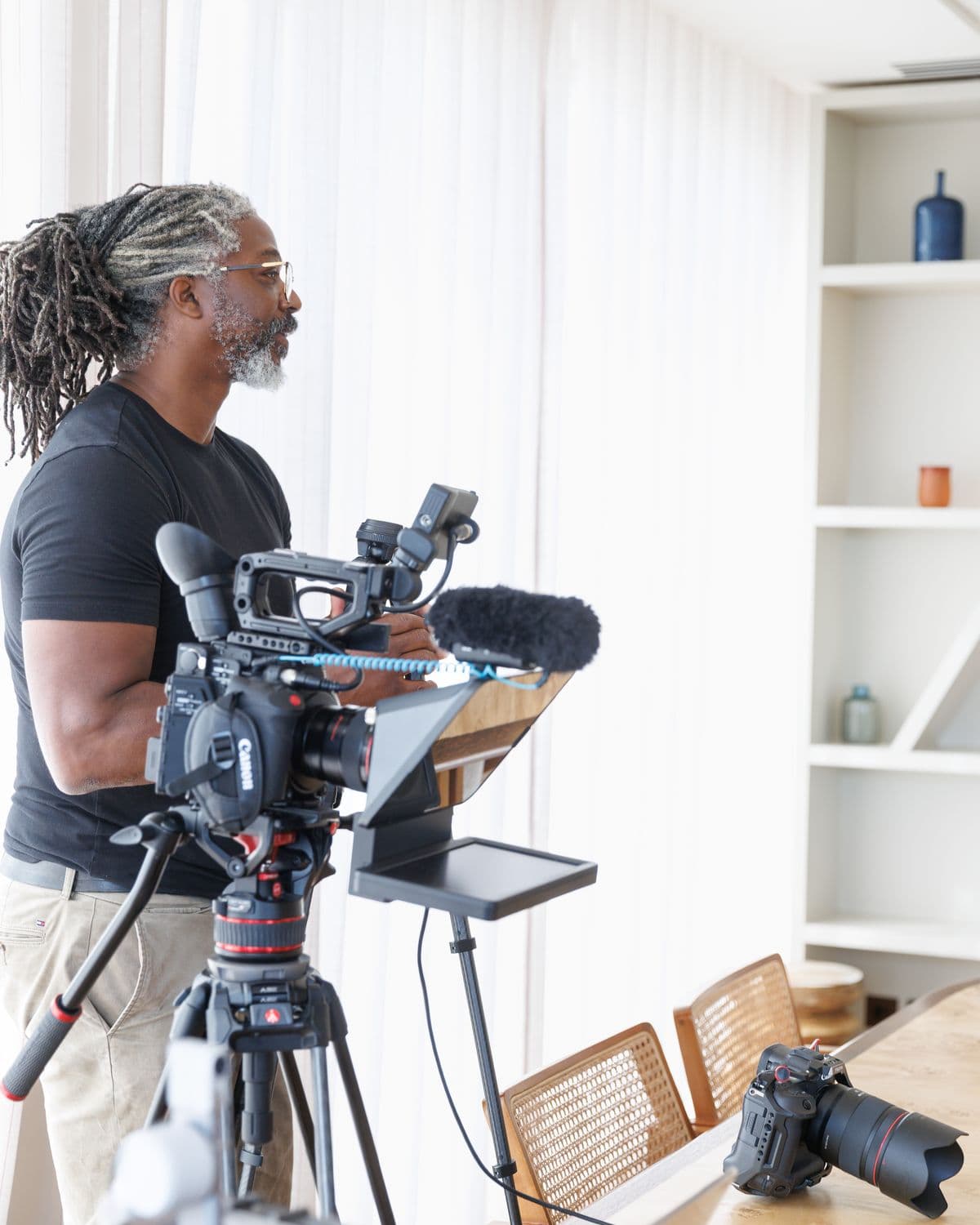 Kevan beside his camera and teleprompter on a tripod, a second camera on the table, looking towards where his subject will sit