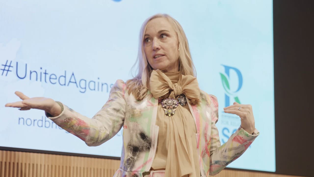 Helene speaking on stage at the conference, one hand held out towards the big screen showing #UnitedAgainst and nordbridgesglobal