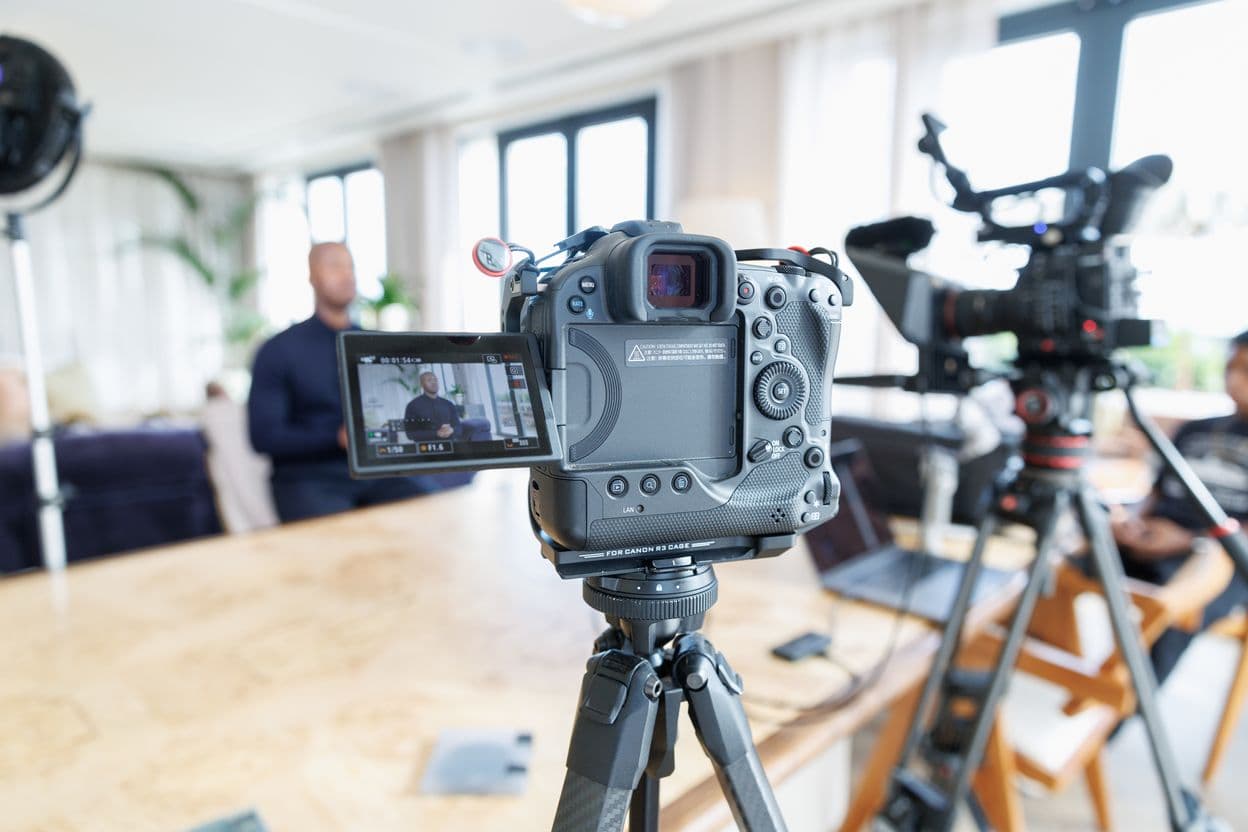 A camera on a tripod in the foreground, its screen showing Undray Griffith seated at the far end of the room, with a second camera behind it, on the August 2025 filming day